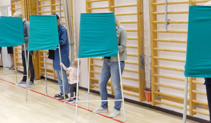 Voters standing inside green fabric voting booths lined up in a gymnasium.