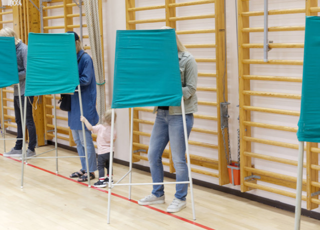 Voters standing inside green fabric voting booths lined up in a gymnasium.