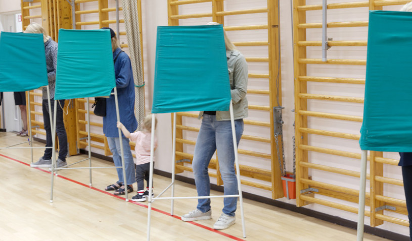 Voters standing inside green fabric voting booths lined up in a gymnasium.