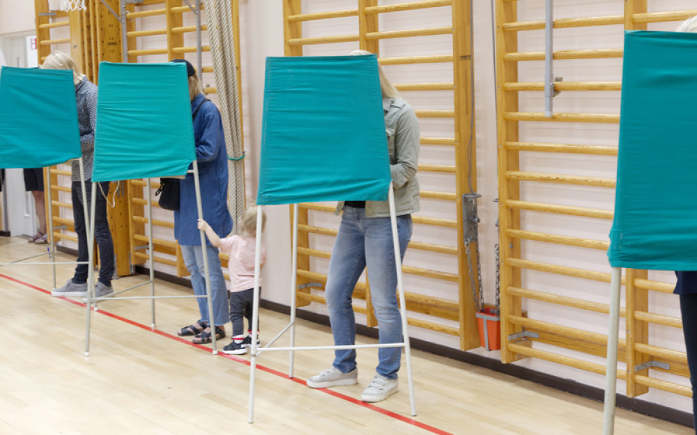 Voters standing inside green fabric voting booths lined up in a gymnasium.