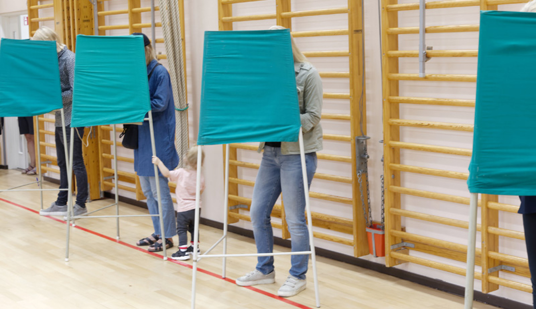 Voters standing inside green fabric voting booths lined up in a gymnasium.