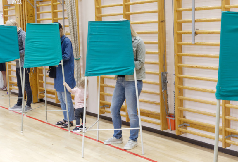 Voters standing inside green fabric voting booths lined up in a gymnasium.