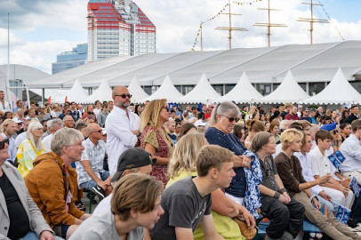 A large crowd seated outdoors at a public event with white tents and a red-and-white building in the background.