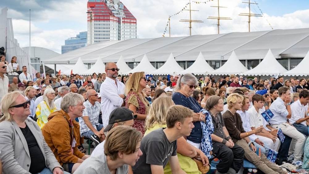 A large crowd seated outdoors at a public event with white tents and a red-and-white building in the background.