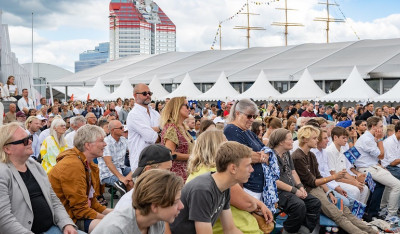 A large crowd seated outdoors at a public event with white tents and a red-and-white building in the background.