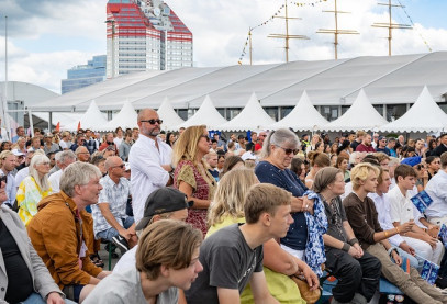 A large crowd seated outdoors at a public event with white tents and a red-and-white building in the background.
