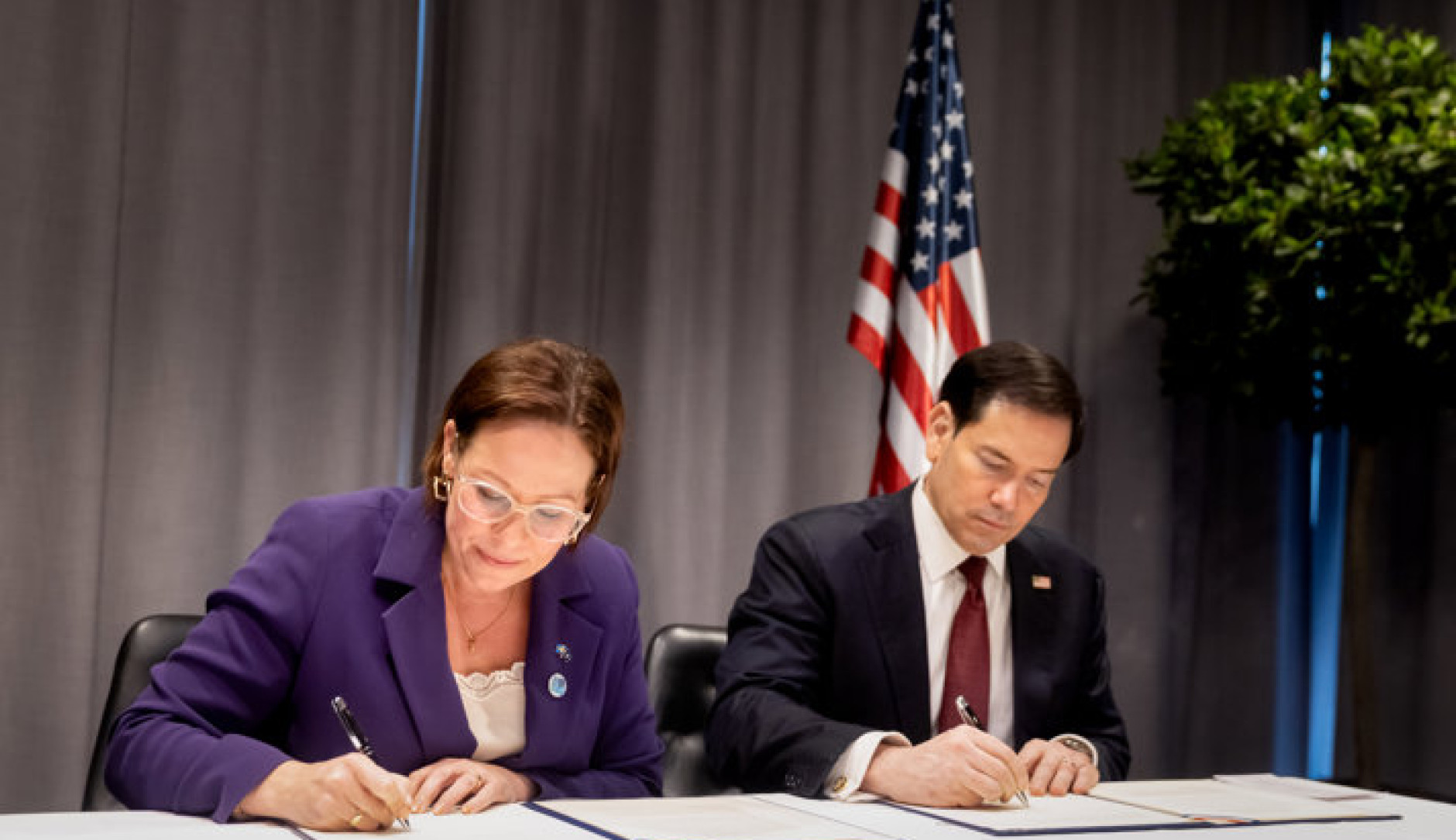 Maria Malmer Stenergard and Marco Rubio sitting at a table, signing official documents in front of an American flag.