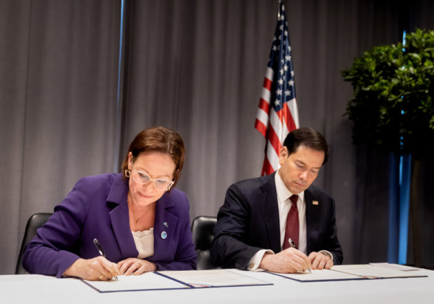 A woman in a purple blazer and a man in a dark suit sitting at a table, signing official documents in front of an American flag.