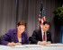 A woman in a purple blazer and a man in a dark suit sitting at a table, signing official documents in front of an American flag.