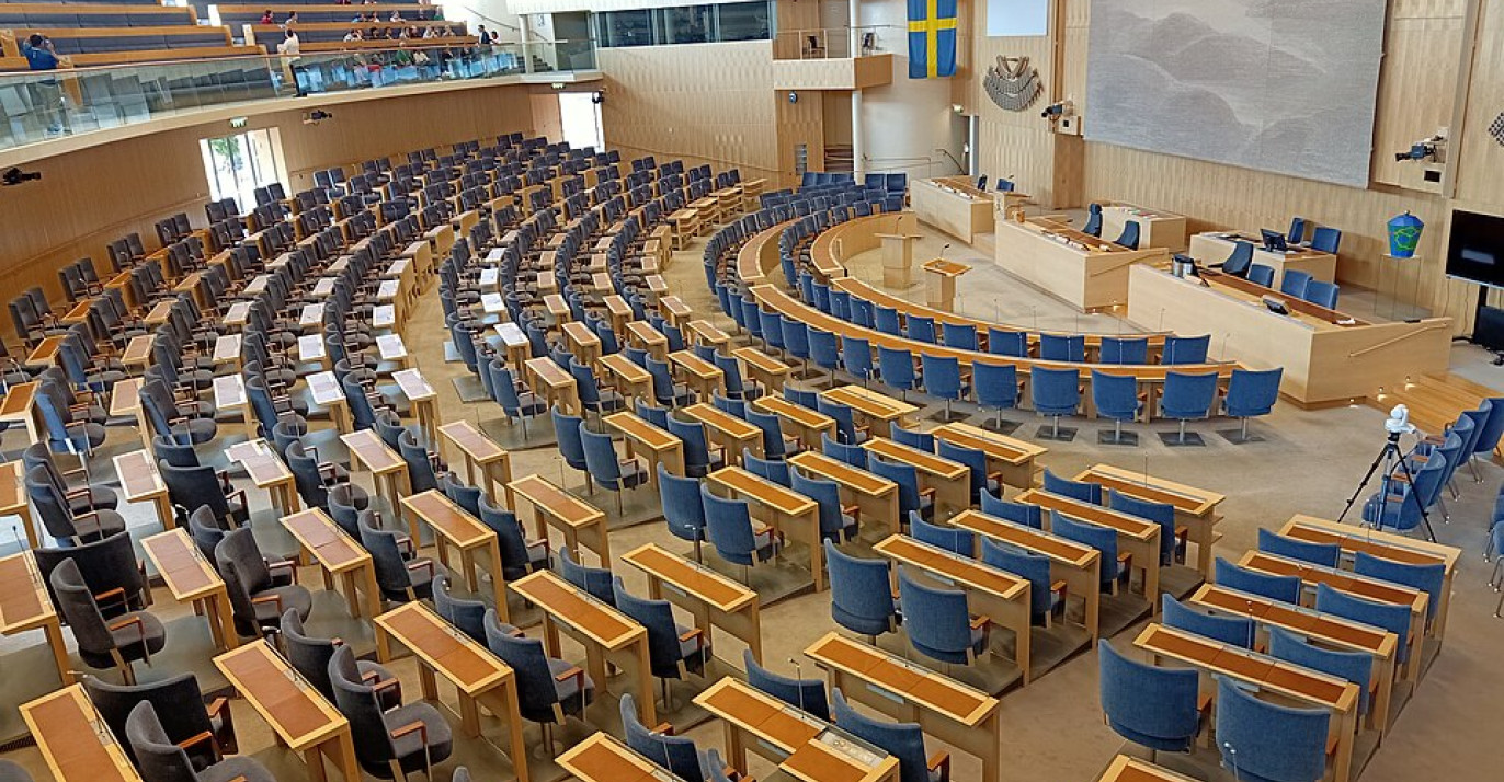 High-angle view of the empty semi-circular plenary chamber of the Swedish Parliament, showing rows of blue chairs and a Swedish flag.