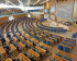 High-angle view of the empty semi-circular plenary chamber of the Swedish Parliament, showing rows of blue chairs and a Swedish flag.