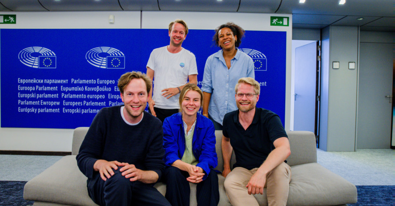 Five people posing on and behind a couch in front of a blue wall featuring the European Parliament logo.
