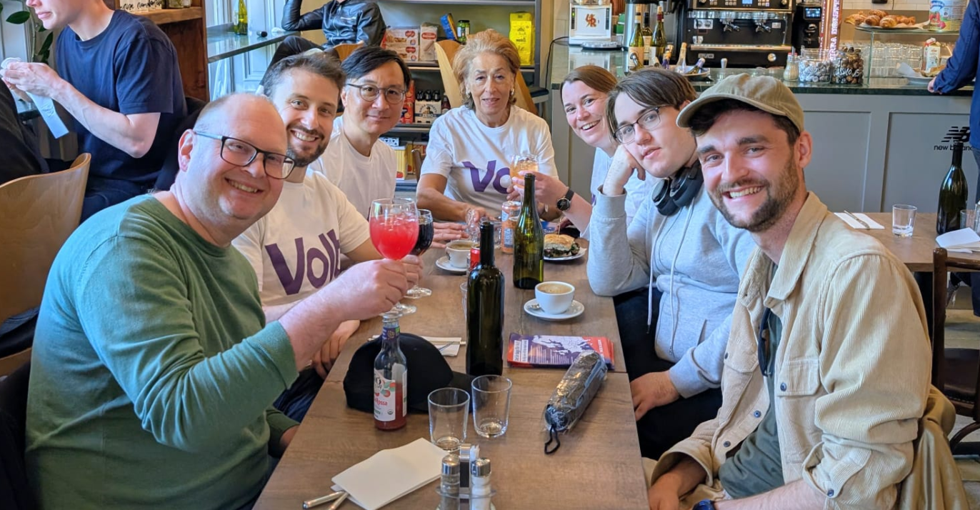 A group of seven people, some wearing white Volt t-shirts, smiling together at a long cafe table with drinks.