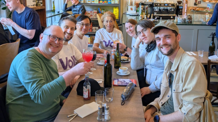 A group of seven people, some wearing white Volt t-shirts, smiling together at a long cafe table with drinks.
